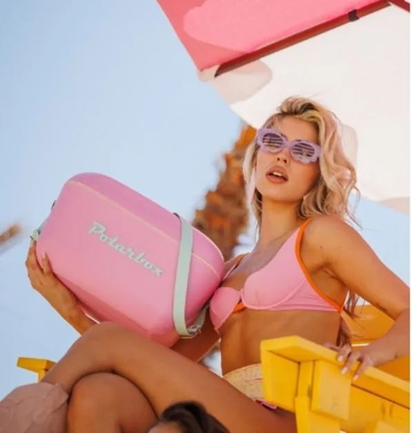 Una donna in bikini rosa tiene una borsa frigo rosa vicino a una sedia gialla.