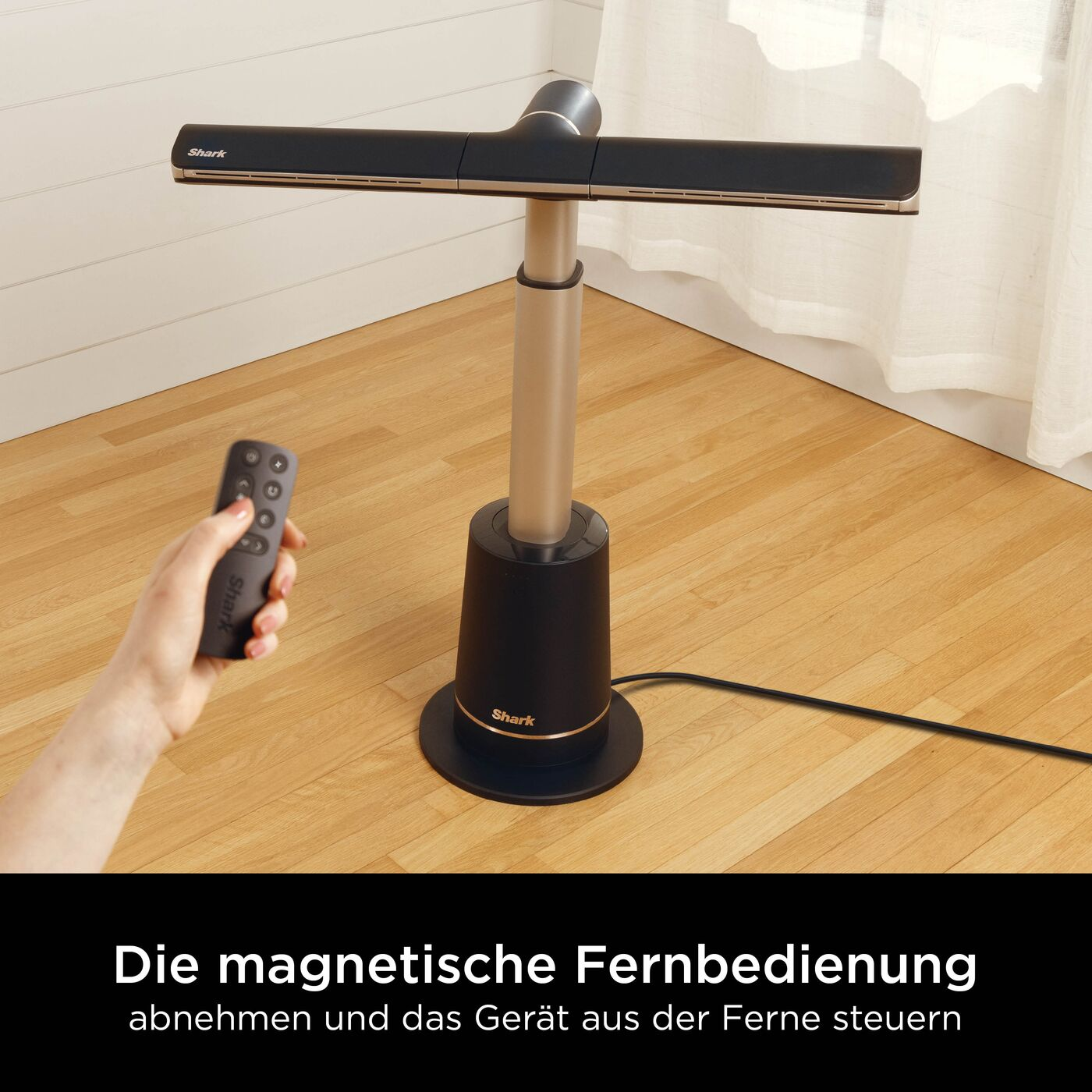 Une main tient une télécommande à côté d'un ventilateur Shark noir sur un parquet.