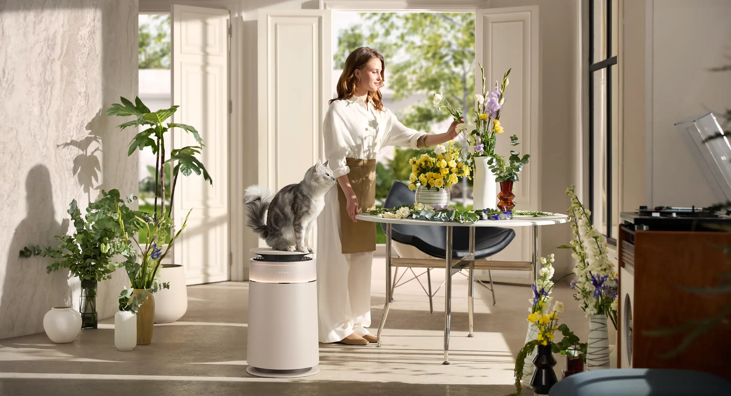 Une femme arrange des fleurs près d'un chat sur un purificateur d'air. Une table avec des fleurs et plantes.
