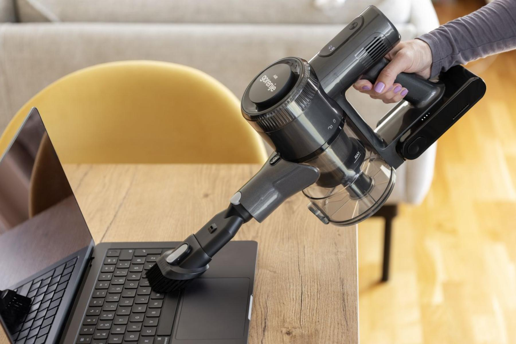 Dłoń trzyma ciemny odkurzacz ręczny, czyszczący klawiaturę laptopa.