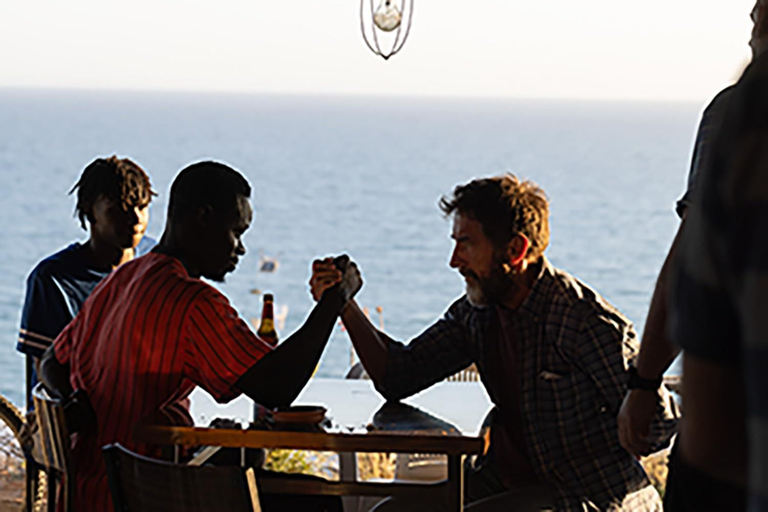 Zwei Männer ringen im Freien am Tisch, Meer im Hintergrund.