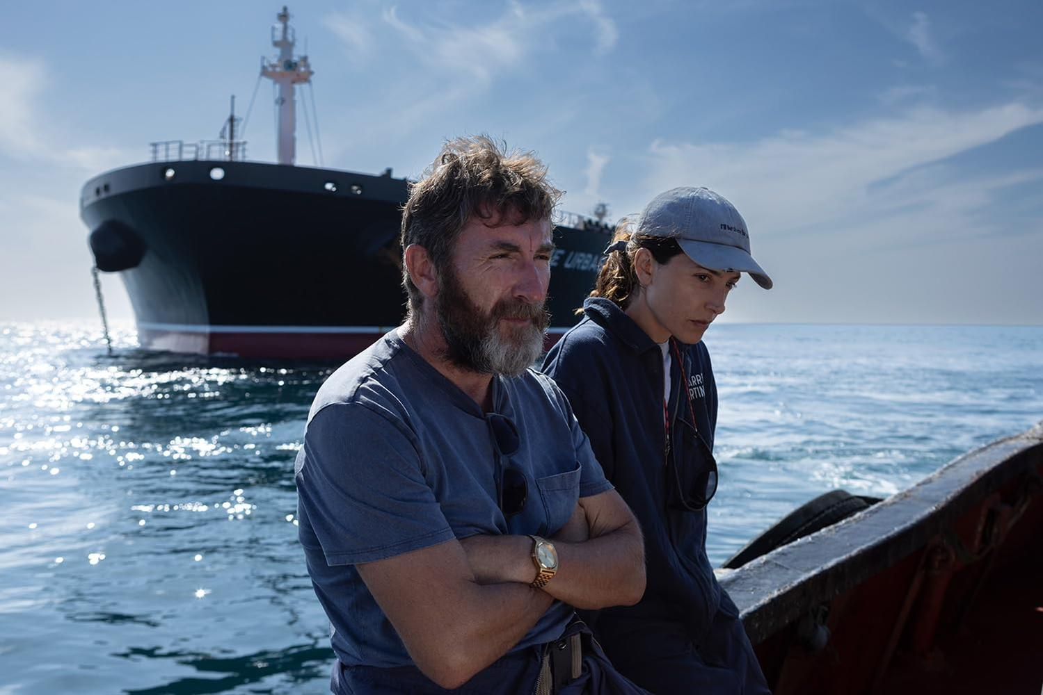 Ein Mann mit Bart und eine Frau mit Kappe sitzen auf einem Boot, ein großes Schiff im Hintergrund.