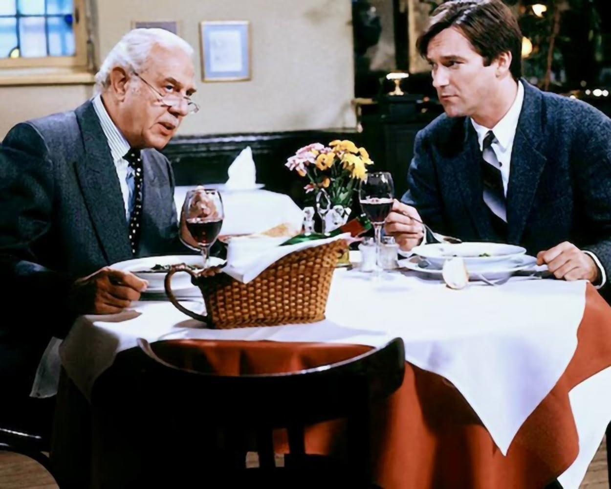 Zwei Männer in Anzügen sitzen an einem Tisch und essen. Ein Mann hält ein Glas Wein.