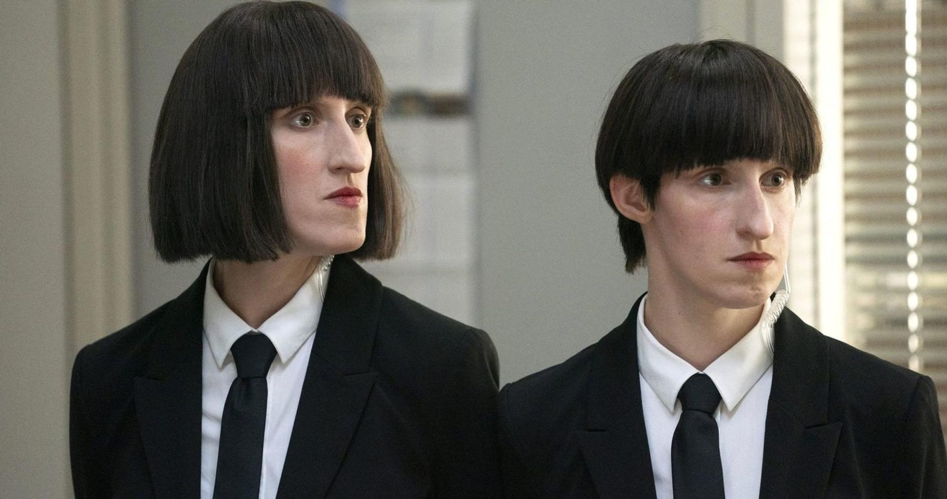 Zwei Personen mit schwarzen Bob-Perücken und Anzügen stehen nebeneinander.