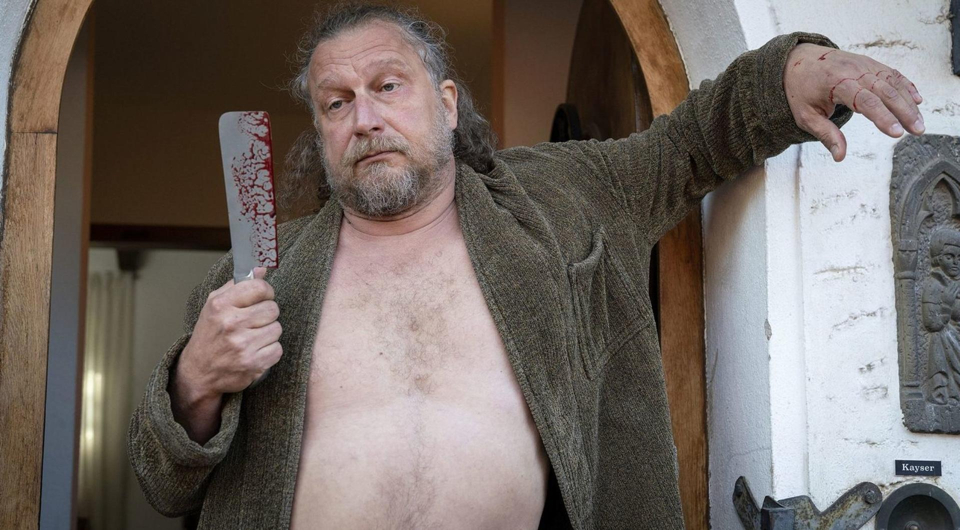Ein Mann mit Bart hält ein blutiges Messer, seine Hand ist verletzt.