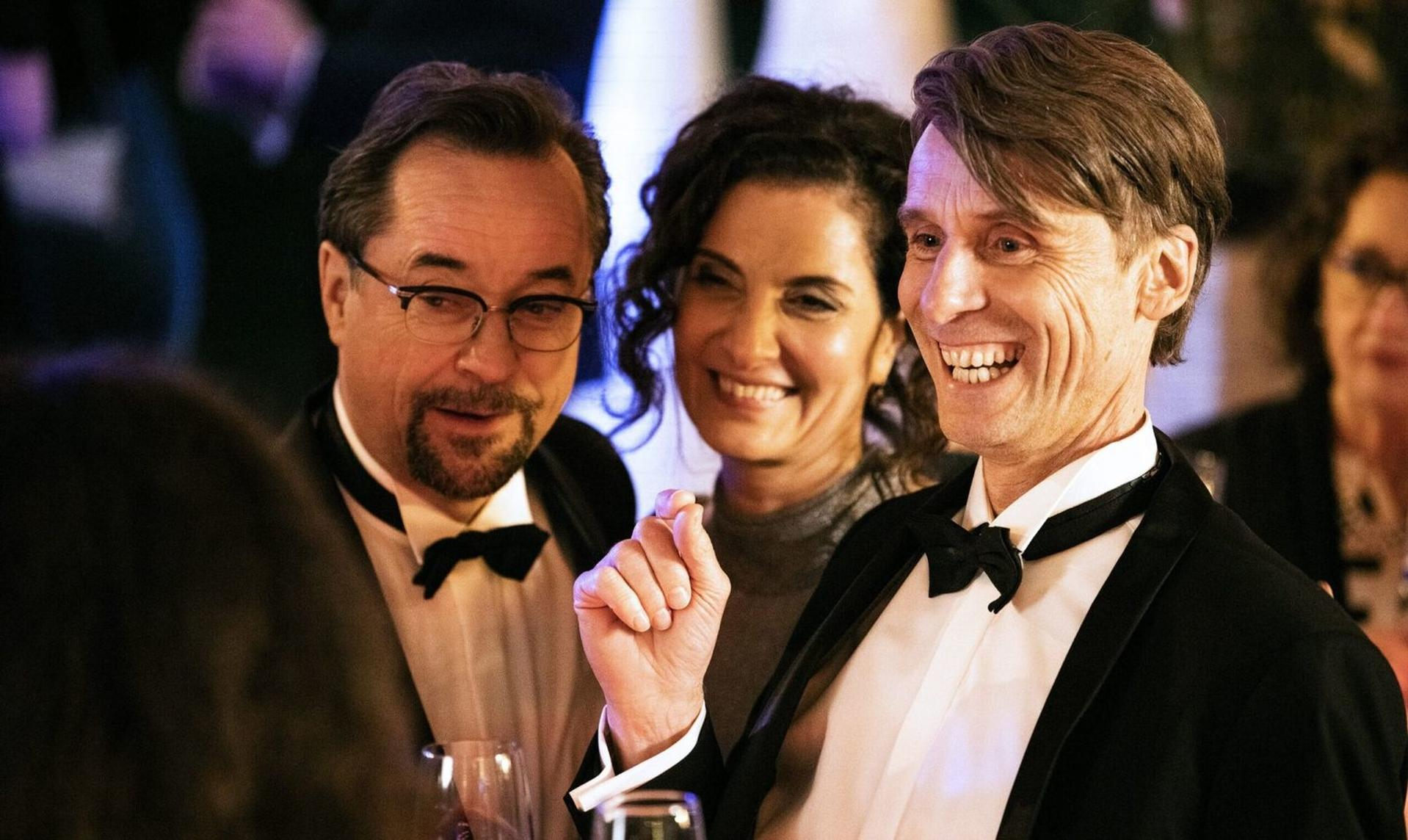 Drei Personen in Anzügen lachen. Der Mann vorne rechts trägt einen Smoking.