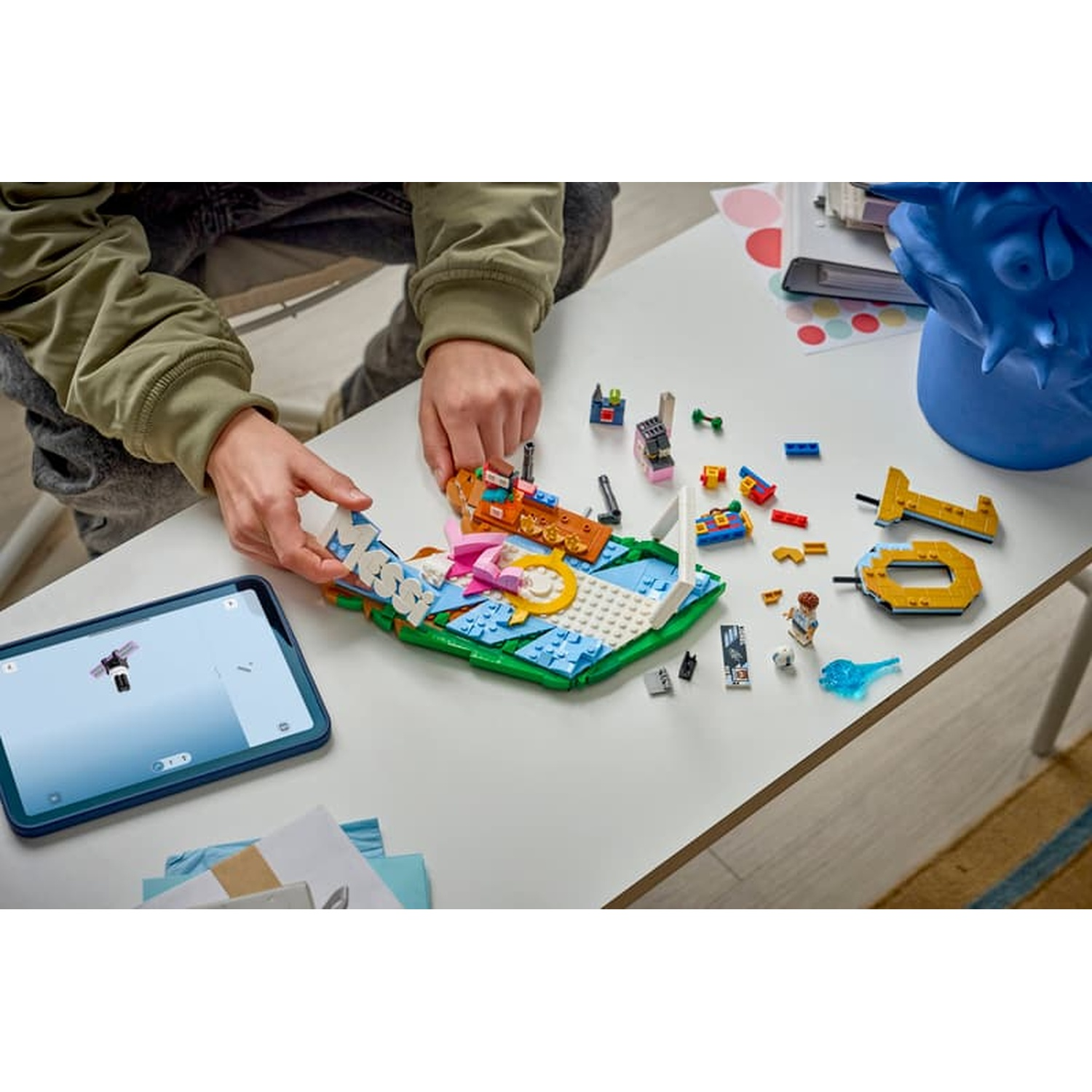 Hands assembling a colorful Lego creation on a white table, with a tablet showing instructions.