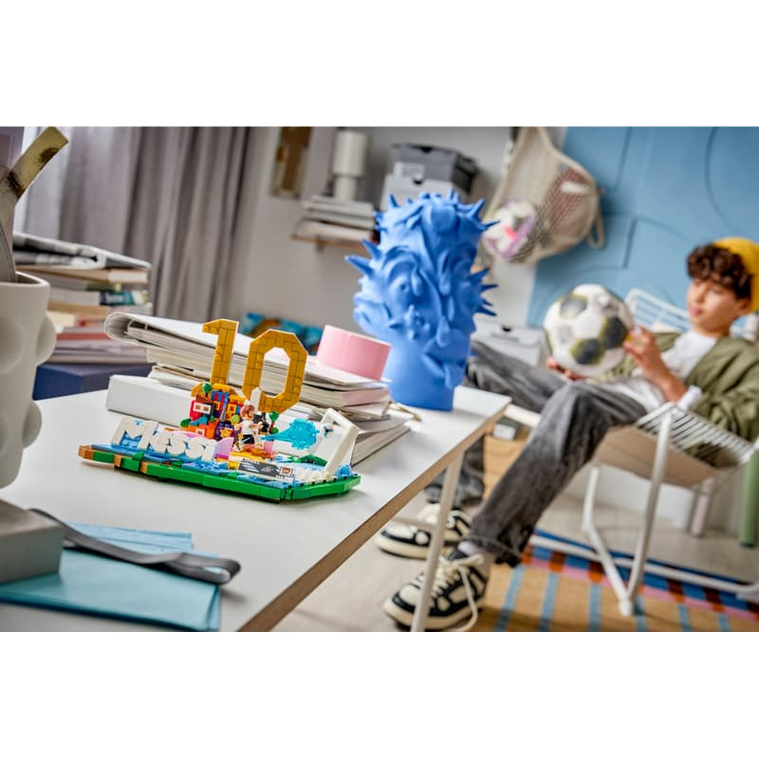 A boy sits holding a soccer ball. A Lego build with the number 10 is on a table.