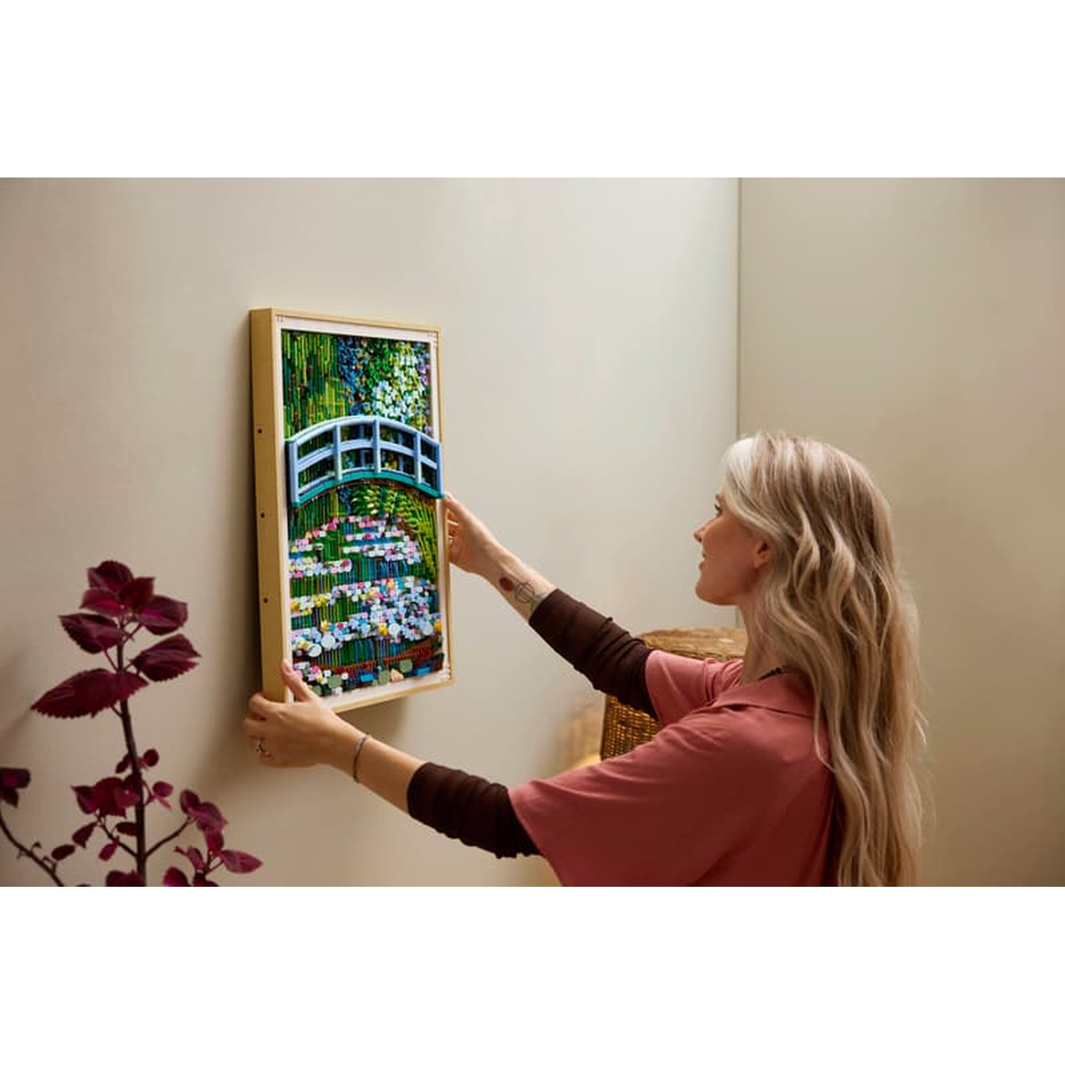 Woman hangs picture of bridge and flowers. Plant on left.
