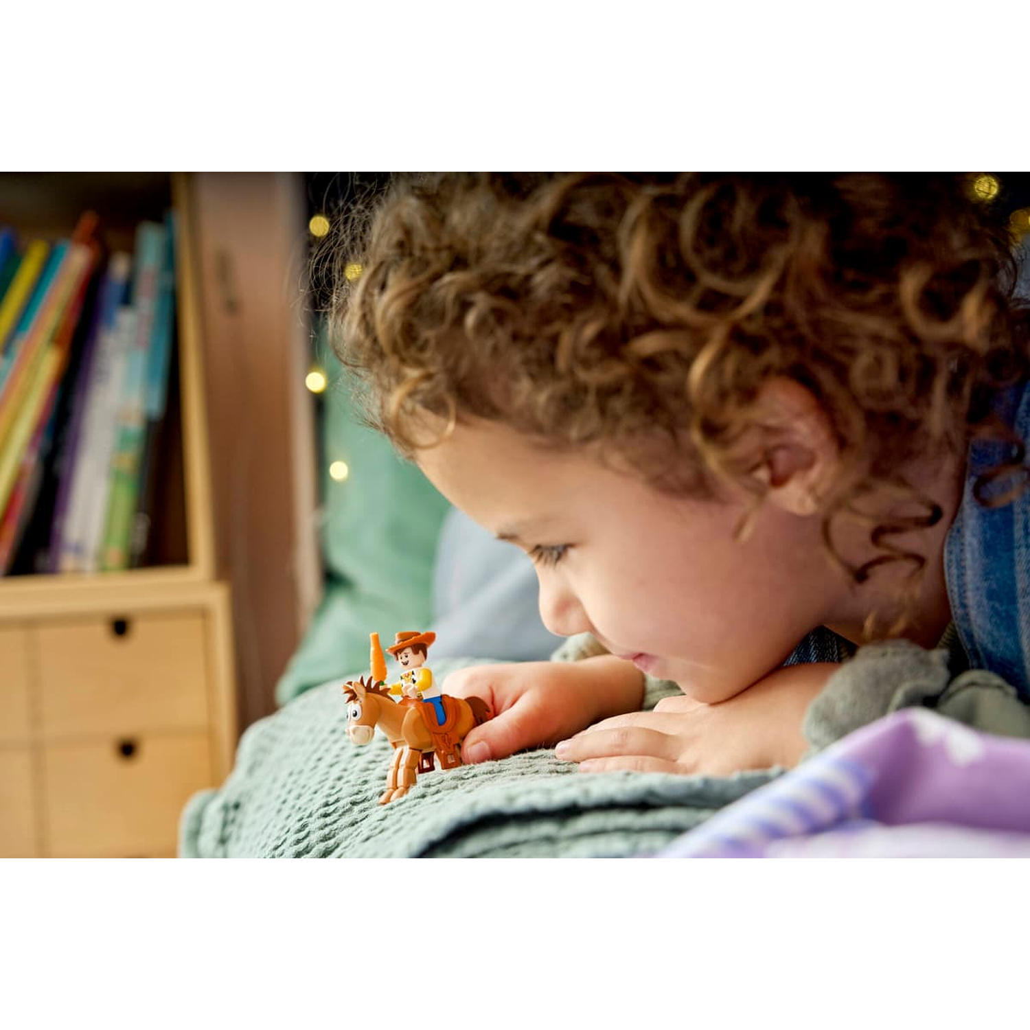 A child plays with a toy cowboy figure riding a brown horse on a green blanket.