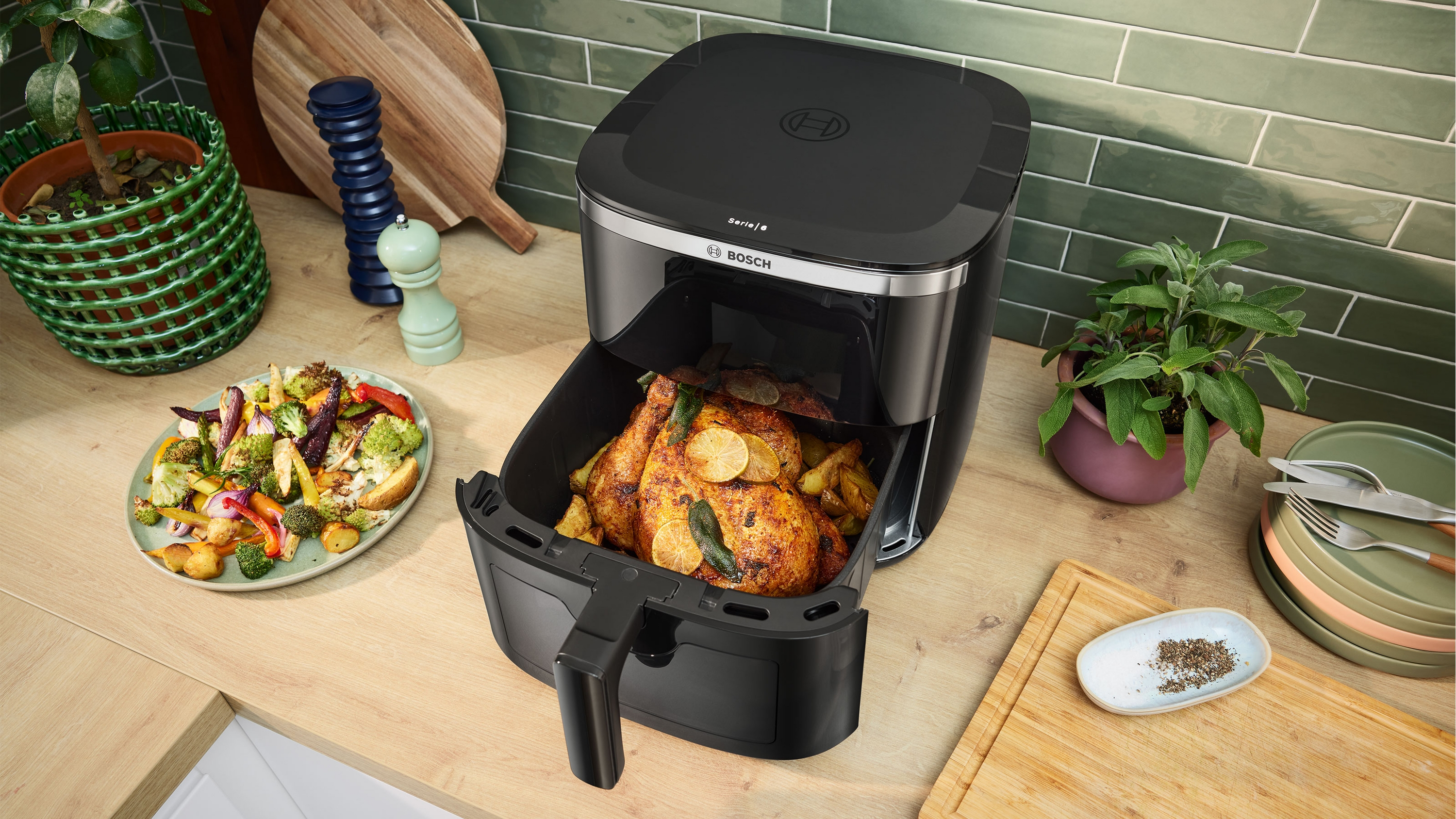 Ein gebratenes Hähnchen mit Zitrone und Kräutern in einem schwarzen Airfryer neben Gemüse.