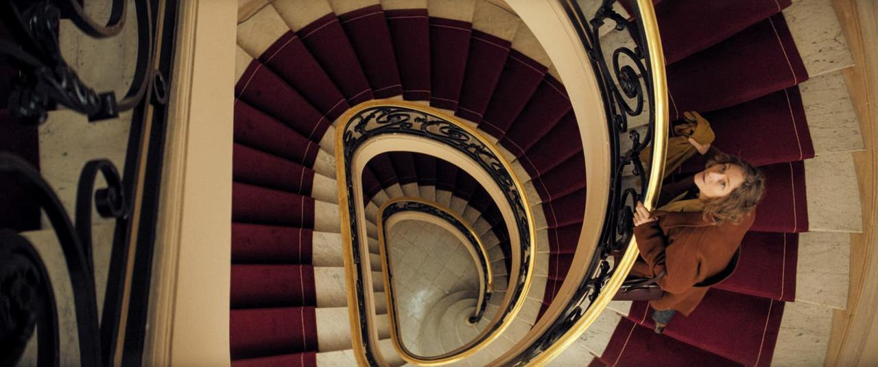 Eine Frau mit braunem Mantel steht auf einer Wendeltreppe mit rotem Teppich.