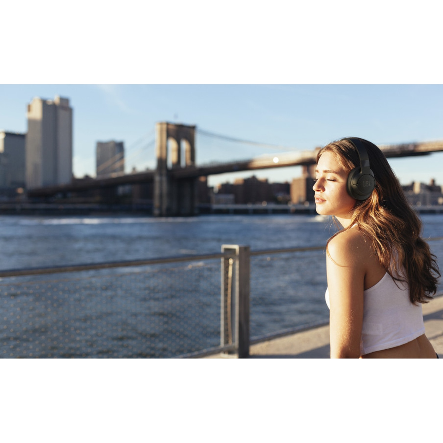 Giovane donna con cuffie seduta vicino all'acqua, con il ponte di Brooklyn sullo sfondo.