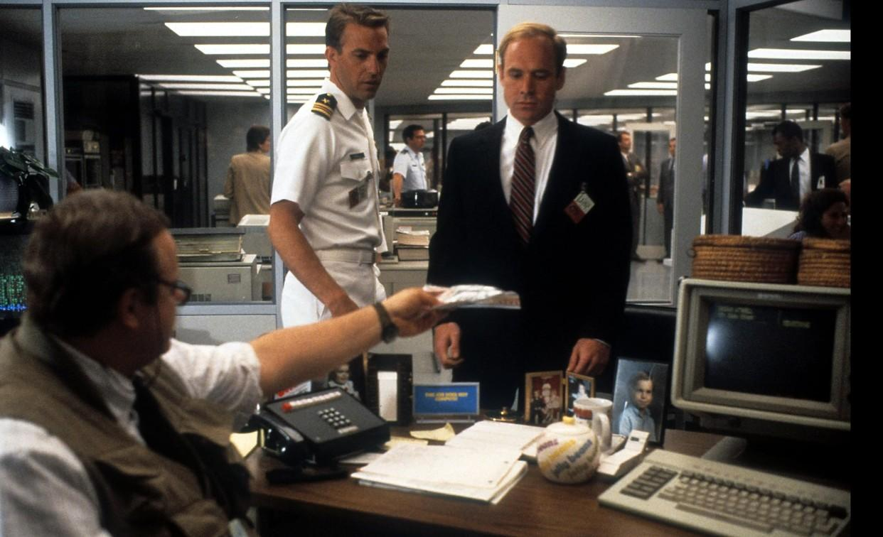 Drei Männer in einem Büro; einer reicht dem anderen ein Bündel Geld.