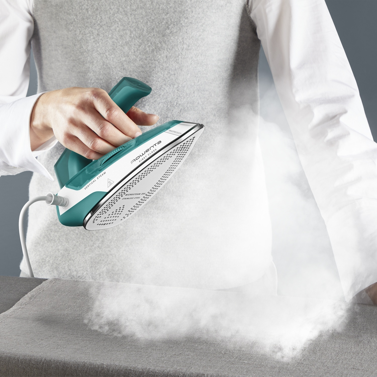 Une main tient un fer turquoise et blanc, émettant de la vapeur sur un tissu gris.