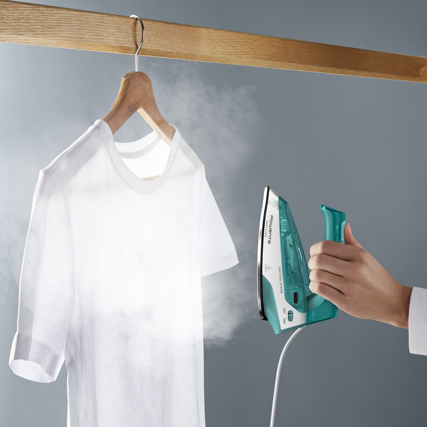 Une main utilise un fer à vapeur bleu sarcelle pour défroisser un t-shirt blanc sur cintre.