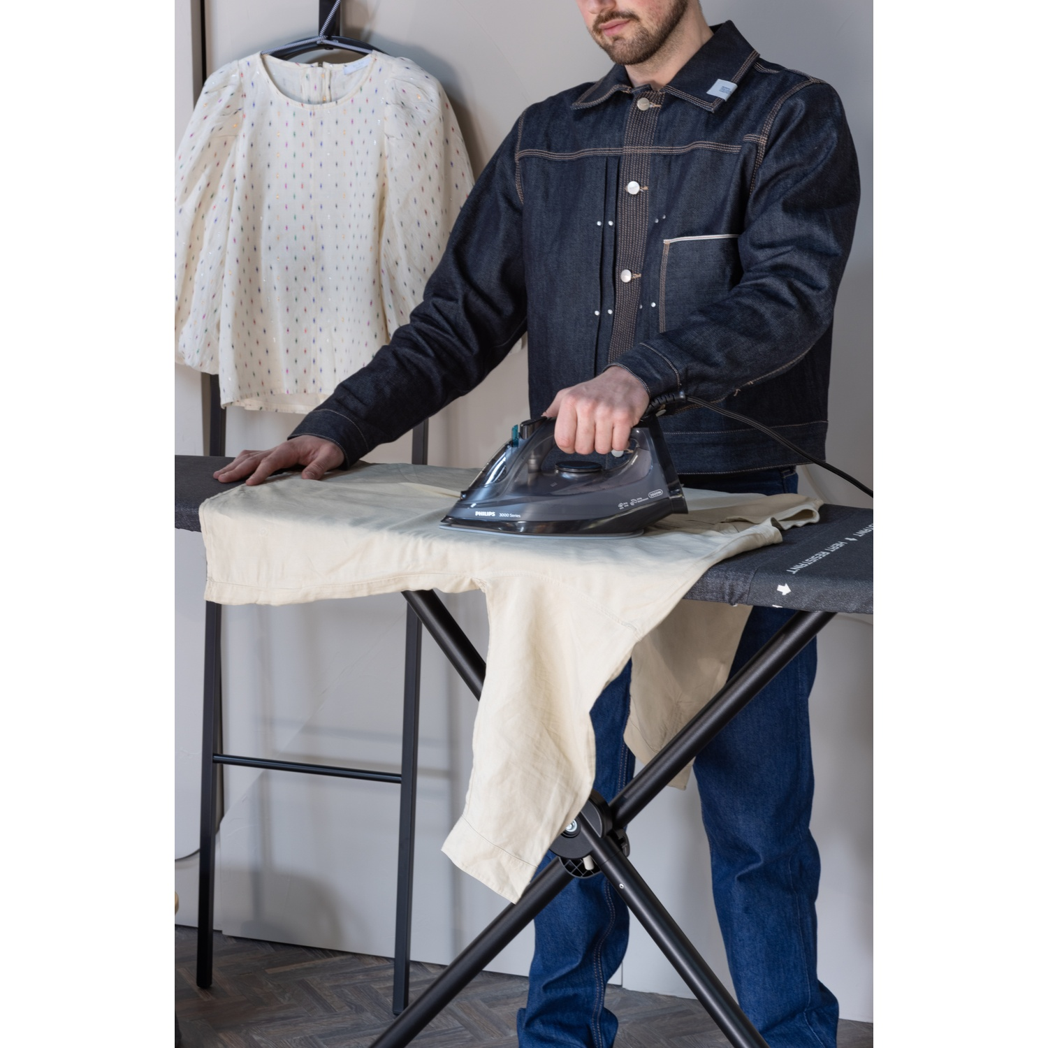 Man in spijkerjack strijkt licht hemd op strijkplank. Witte blouse hangt ernaast.