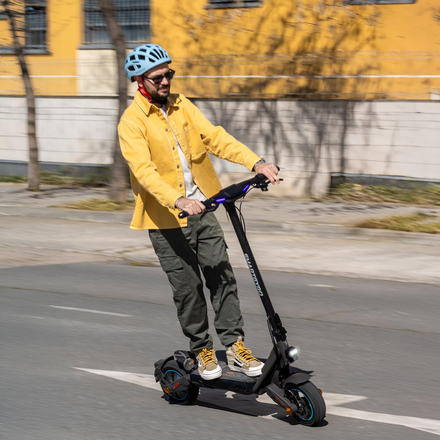 Hombre con camisa amarilla monta patinete eléctrico negro en carretera, con casco azul.