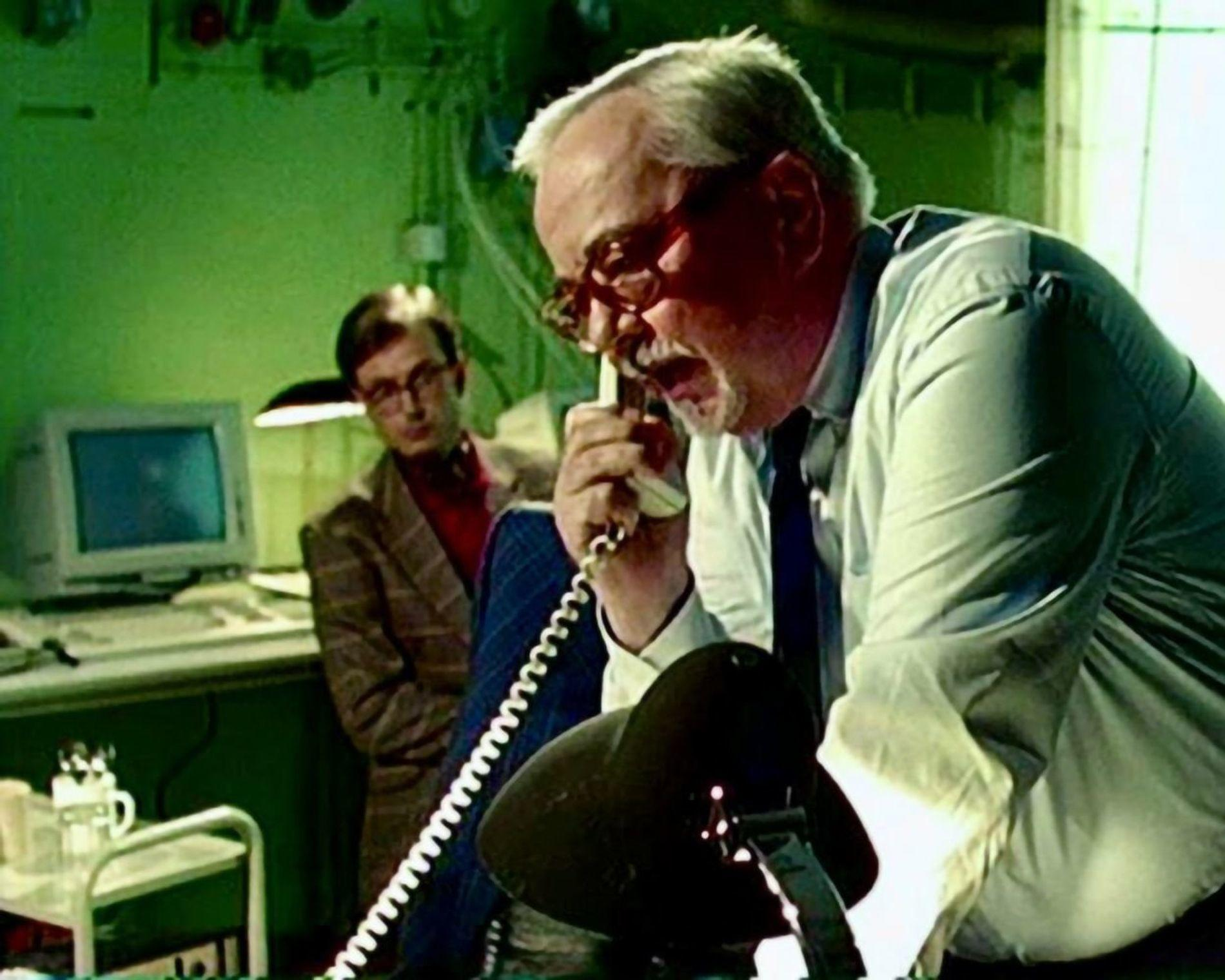 Mann mit Brille telefoniert, ein anderer Mann im Hintergrund sitzt am Schreibtisch.
