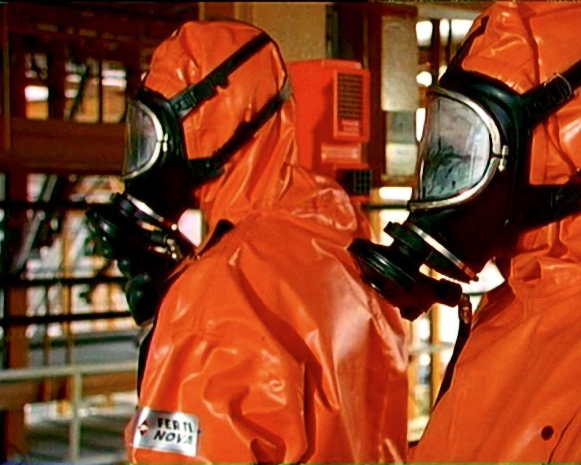 Zwei Personen in orangefarbener Schutzkleidung mit Gasmasken in einem industriellen Umfeld.