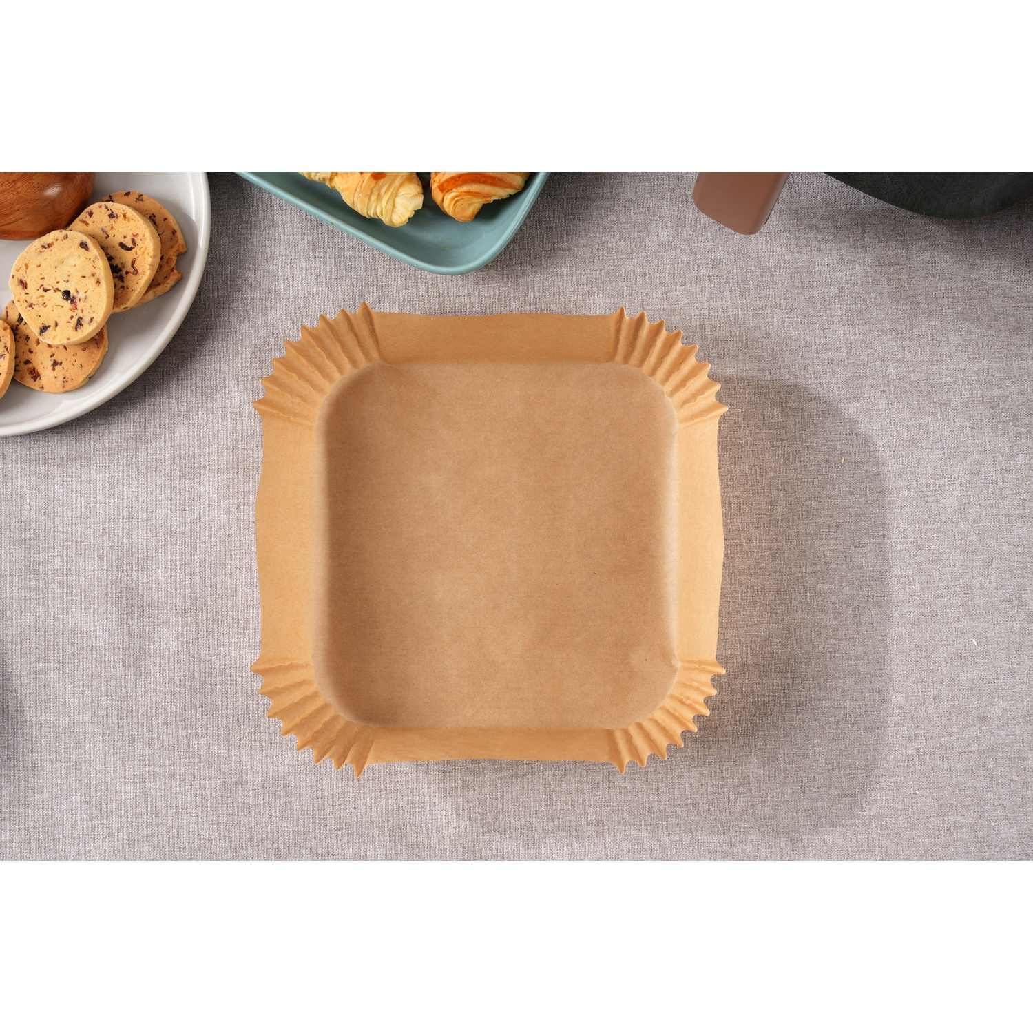 Una forma quadrata di carta da forno marrone con bordo ondulato, accanto a biscotti e croissant.