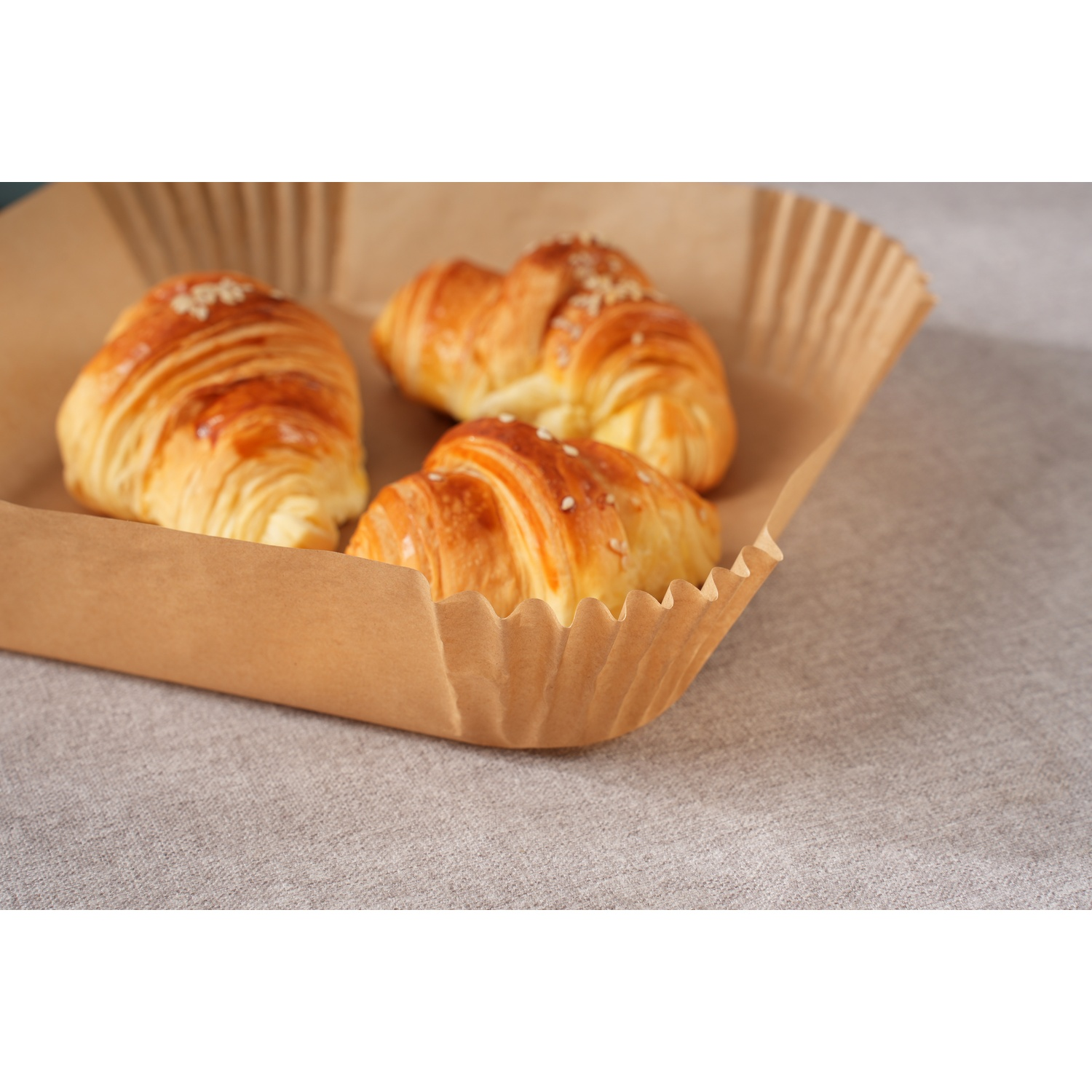 Tre croissant dorati con semi di sesamo in un contenitore di carta marrone su stoffa grigia.