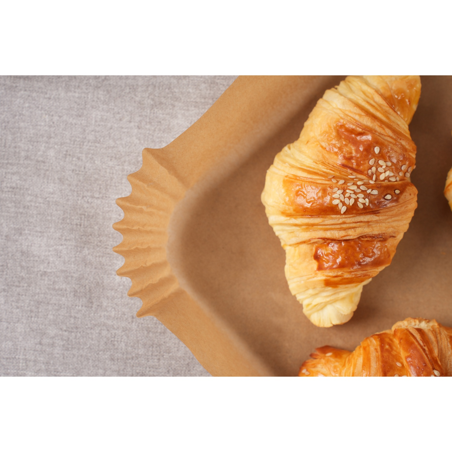 Due croissant dorati con semi di sesamo su carta da forno, su una tovaglia grigia.