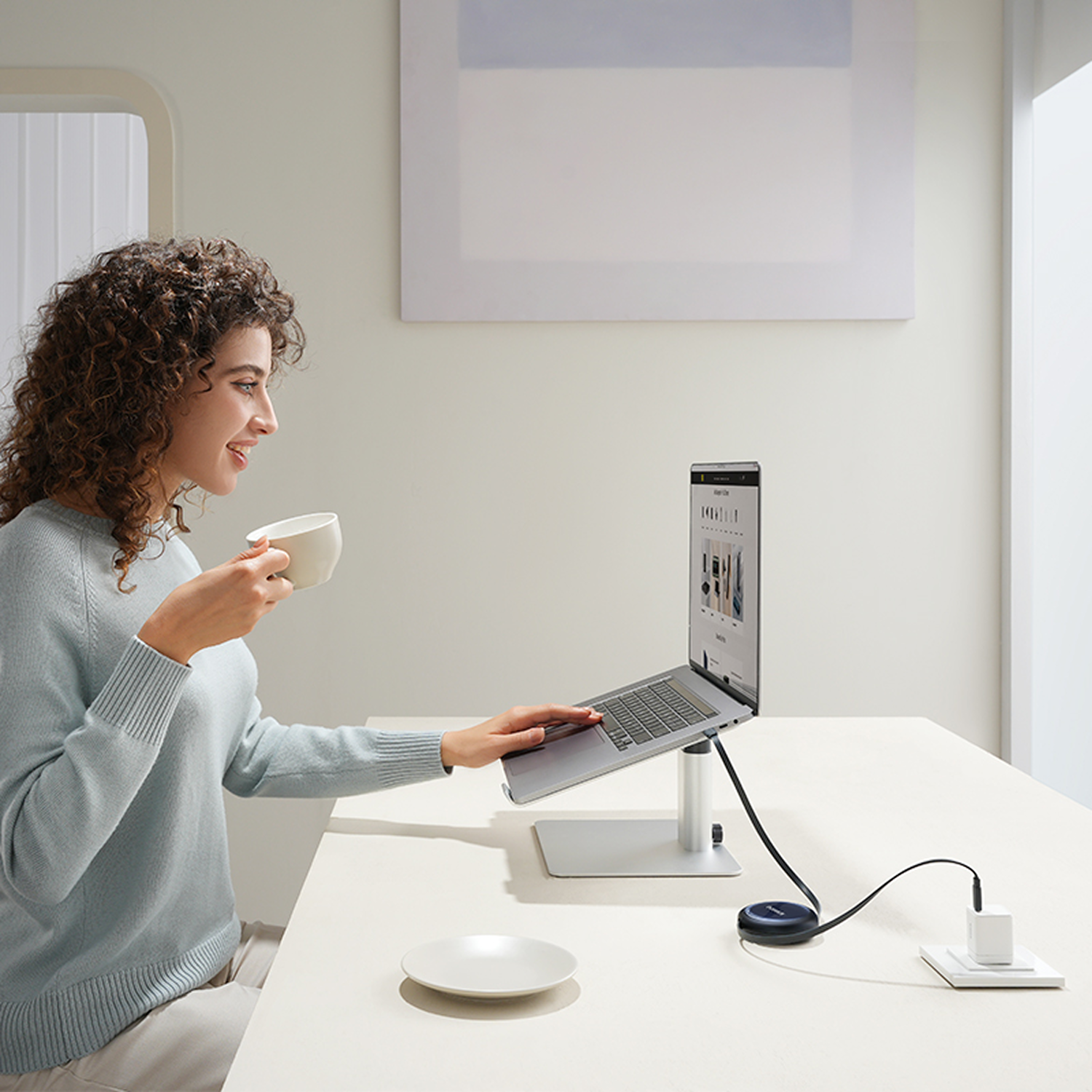 Una mujer con cabello rizado bebe café mientras usa una laptop en un soporte.