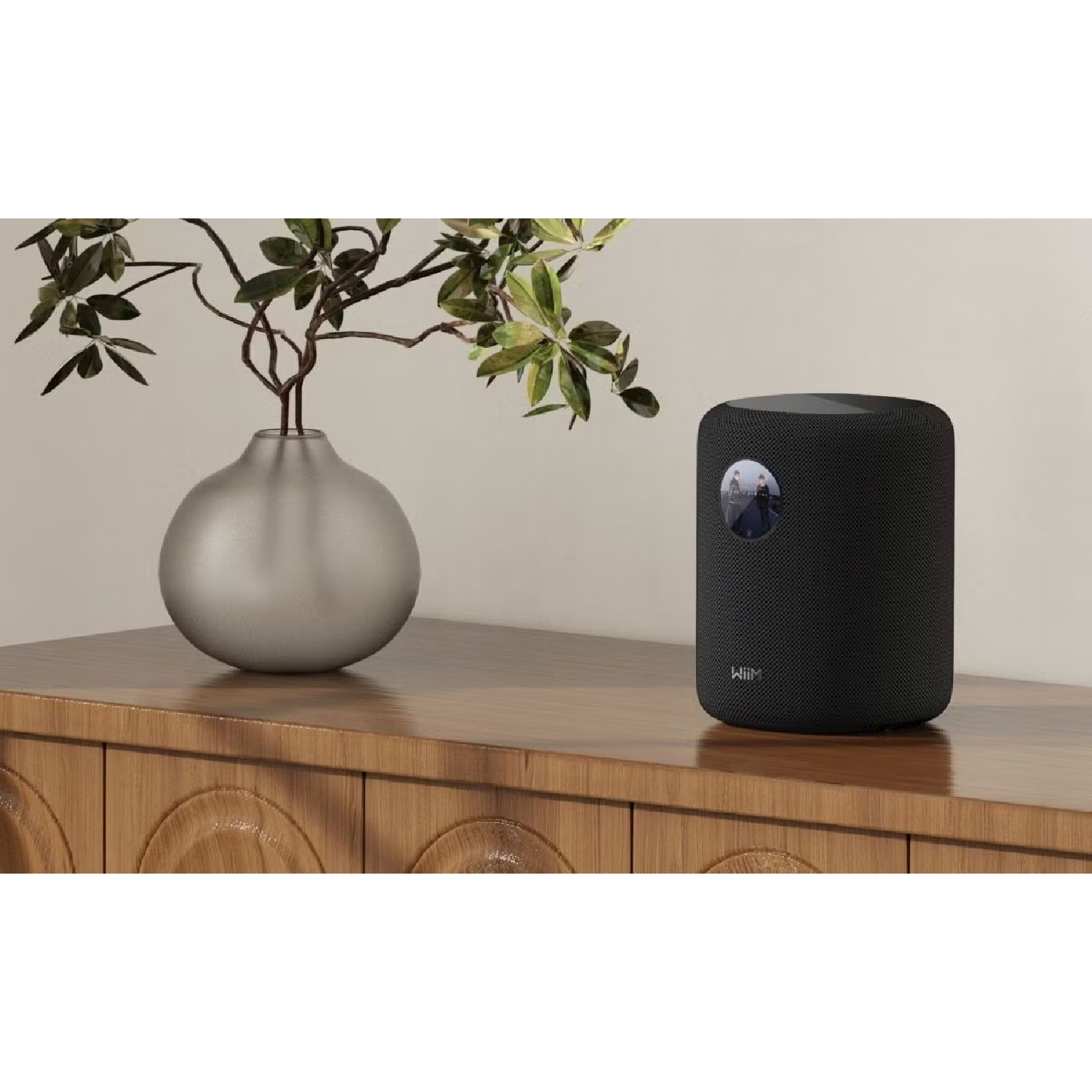 Un altoparlante intelligente nero si trova su una cassettiera di legno accanto a un vaso d'argento con foglie verdi.