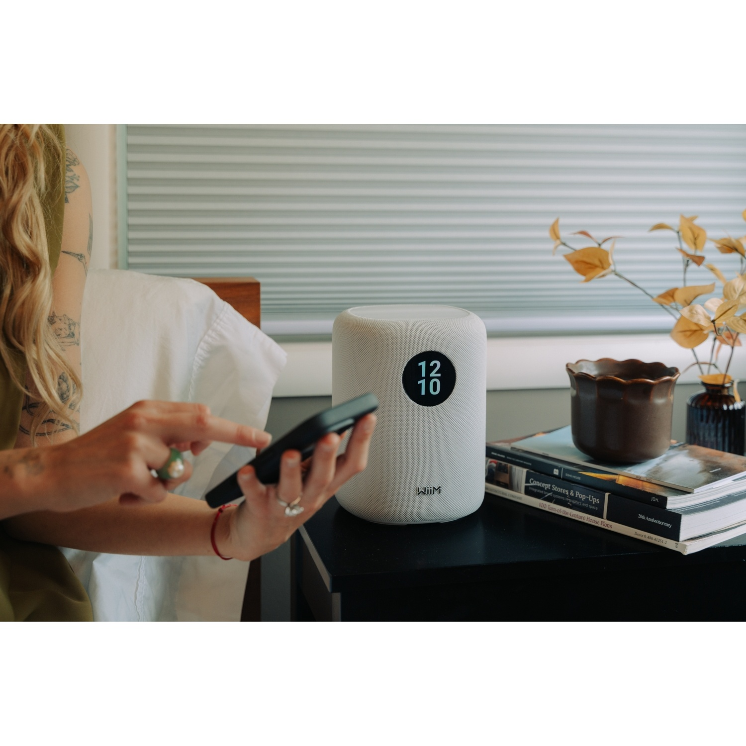 Una persona usa uno smartphone accanto a un altoparlante bianco che mostra le 12:10.