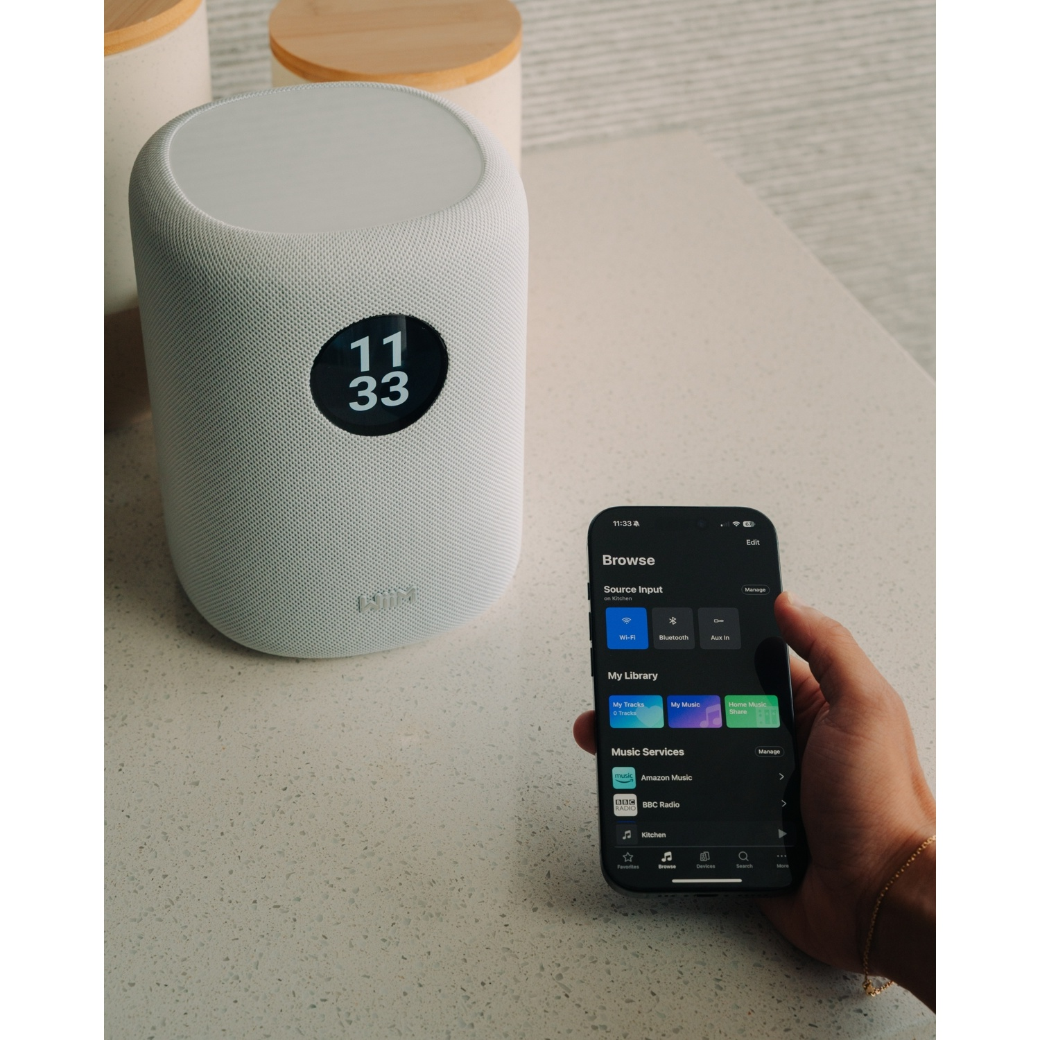 Un altoparlante bianco mostra 11:33 accanto a uno smartphone con un'app musicale.