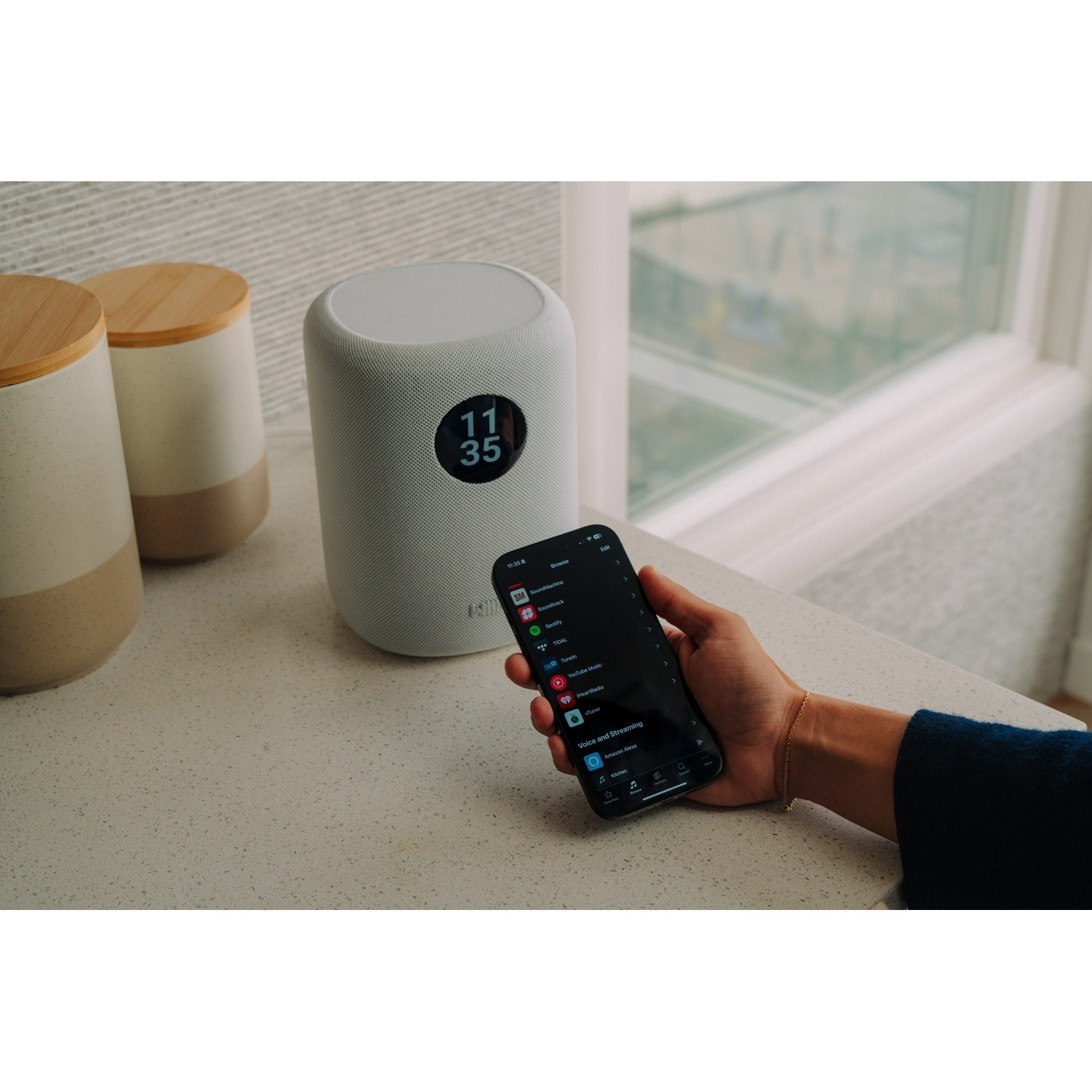 Un altoparlante smart bianco con display dell'orologio e un telefono che mostra un'app.