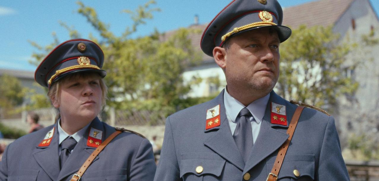 Zwei Männer in grauen Uniformen mit Mützen stehen draußen.