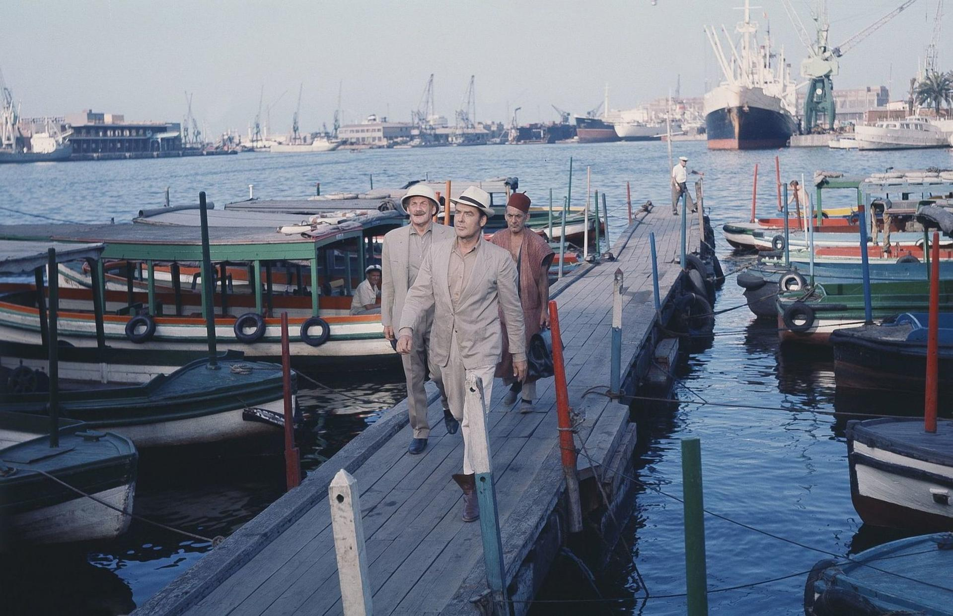 Männer in Hüten gehen auf einem Holzsteg an Booten vorbei, im Hintergrund ein Hafen mit Kränen.