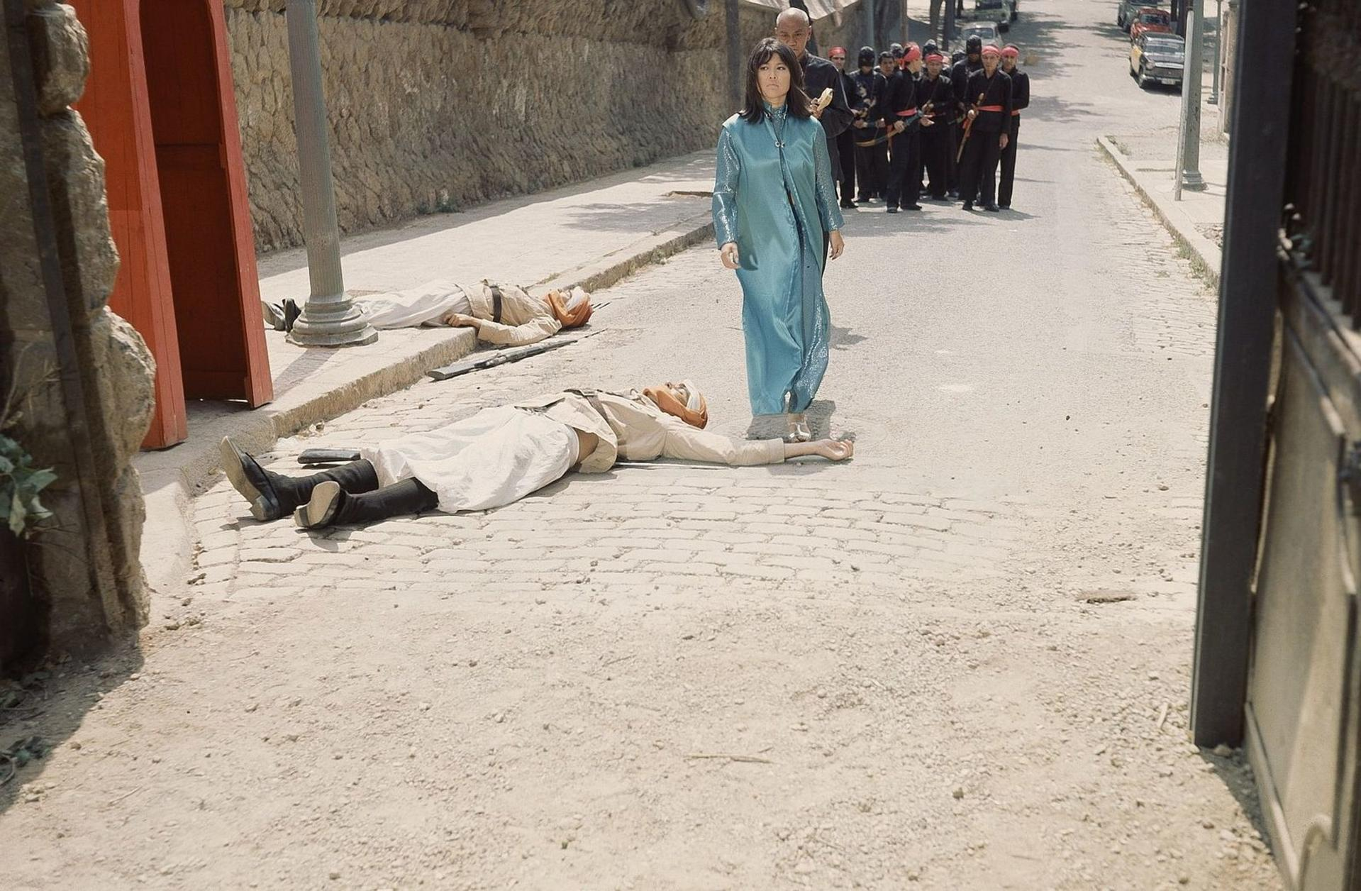 Eine Frau in einem blauen Kleid geht an zwei toten Männern vorbei, eine Gruppe in Schwarz folgt.