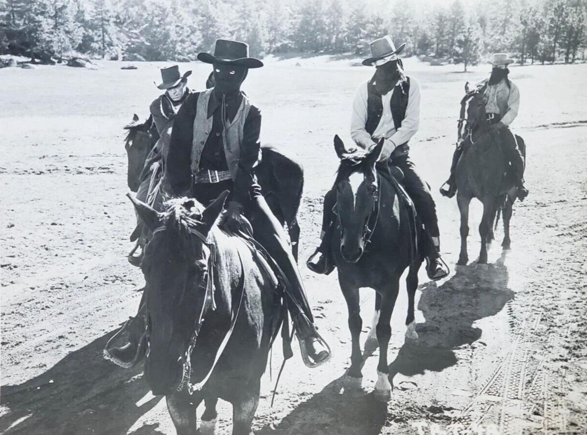 Vier Reiter in Cowboykleidung reiten auf Pferden in einer trockenen, felsigen Landschaft.