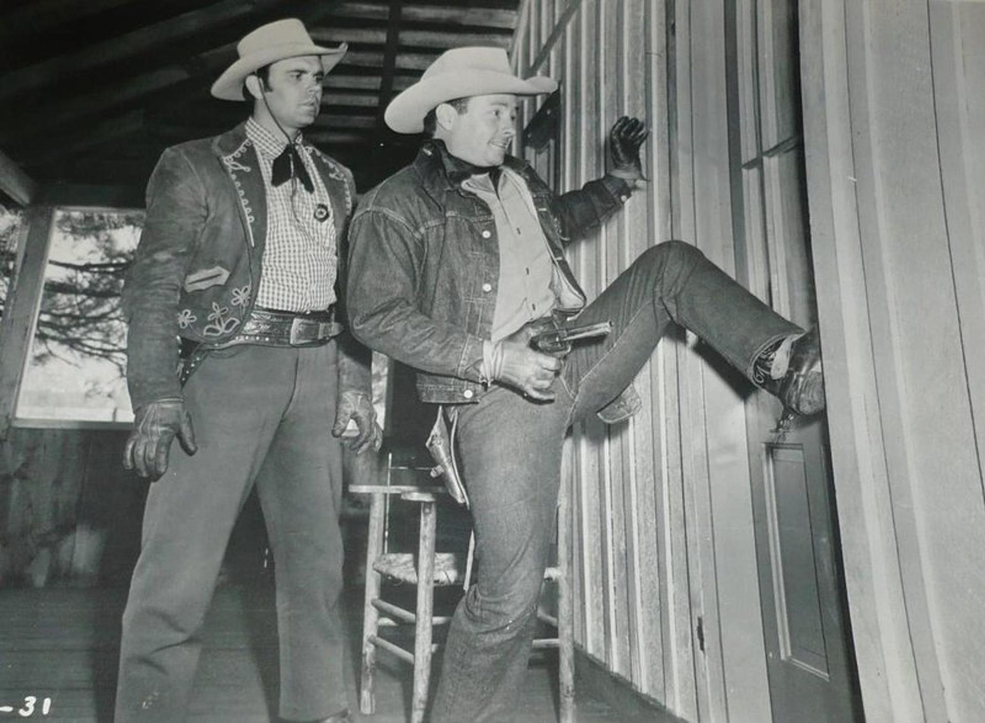 Zwei Cowboys in einem Holzhaus, einer hält eine Pistole, der andere steht daneben.