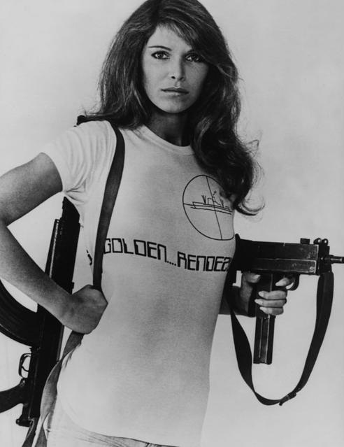 Eine Frau mit langen Haaren hält eine Maschinenpistole und trägt ein T-Shirt mit Aufschrift.