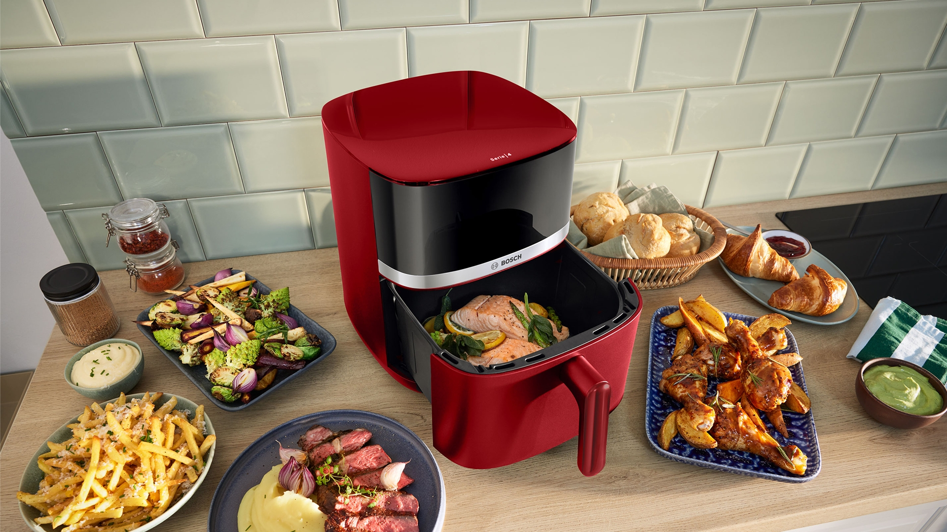 Ein roter Bosch Airfryer mit zubereiteten Speisen wie Lachs, Hähnchen und Beilagen.