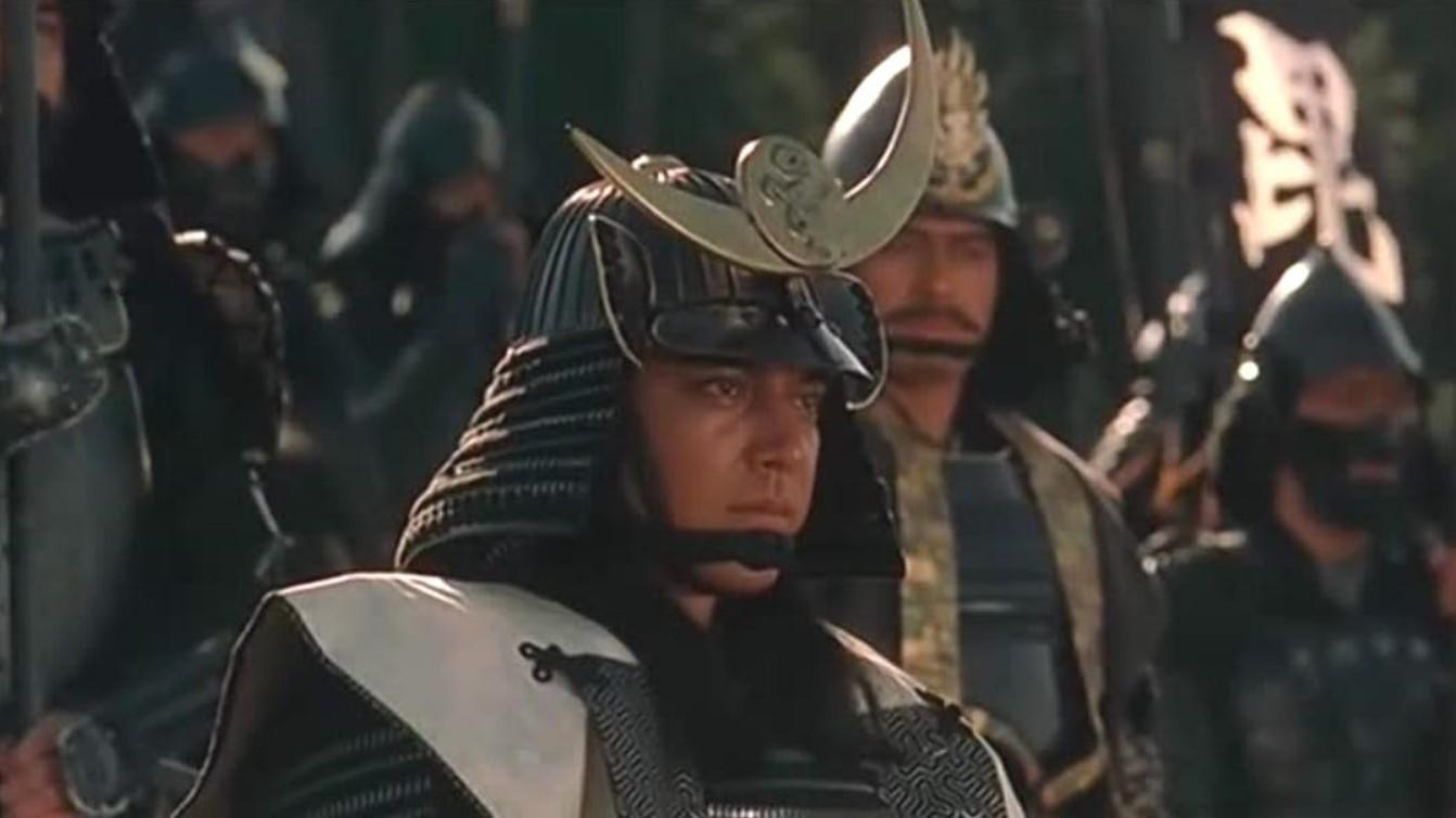 Ein Samurai mit Helm und Rüstung steht vor einer Gruppe anderer Krieger im Freien.