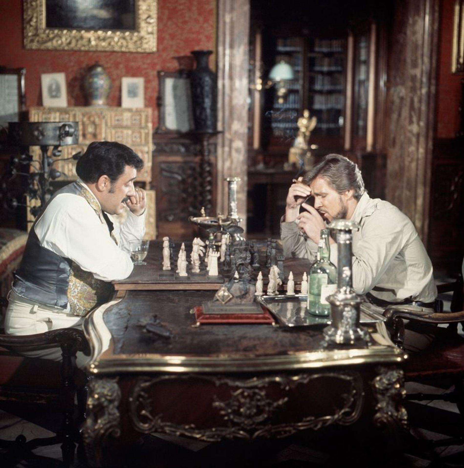 Zwei Männer spielen Schach in einem opulenten Raum. Einer raucht, der andere trinkt.