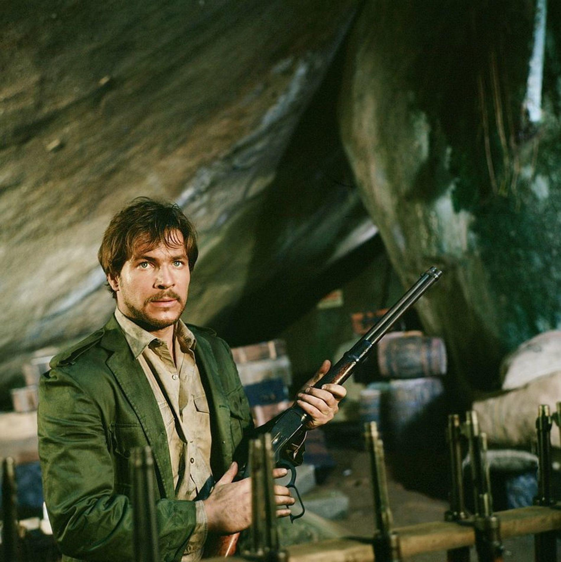 Ein Mann in einem grünen Mantel hält ein Gewehr in einer Höhle mit Fässern im Hintergrund.