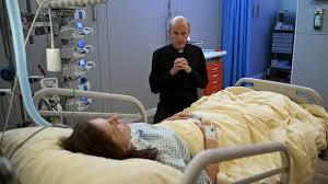 Ein Mann in Schwarz kniet neben einem Bett, auf dem eine Frau liegt. Medizinische Geräte sind sichtbar.