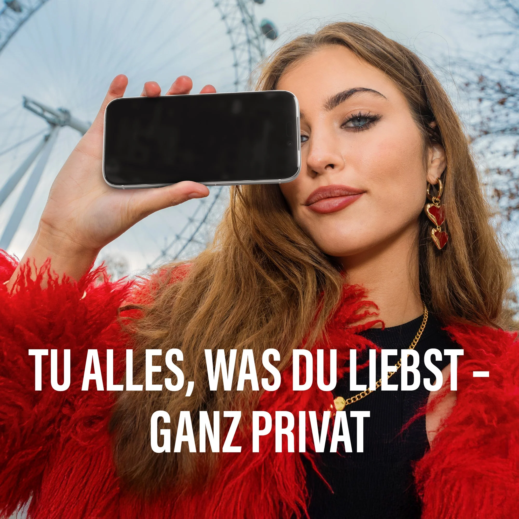 Femme tenant un téléphone devant une grande roue. Manteau rouge, boucles d'oreilles cœur or.