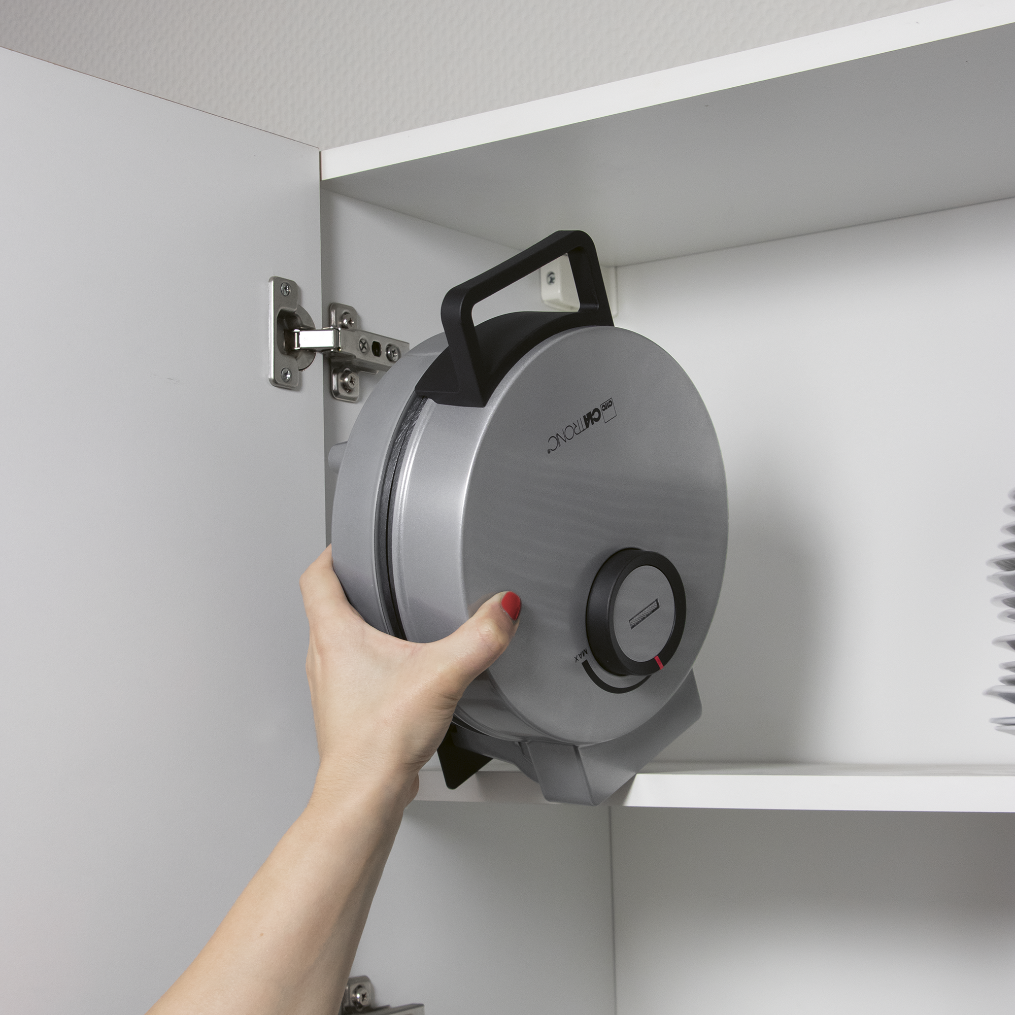Eine Hand nimmt einen silbernen Waffelautomaten aus einem weißen Schrank.