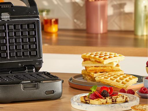 Una piastra per waffle aperta è accanto a una pila di waffle dorati e un piatto con un waffle guarnito.