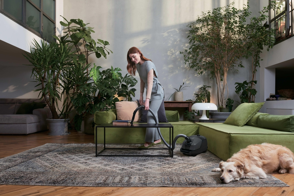 Eine Frau saugt ein Wohnzimmer mit grünen Sofas, Pflanzen und einem Hund.