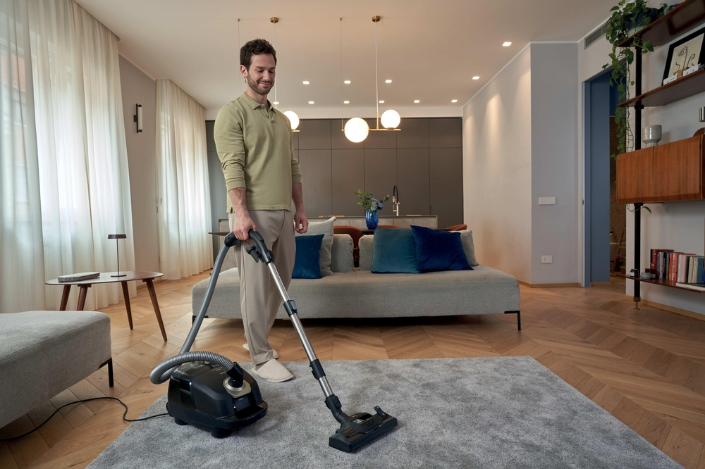 Mann saugt Teppich in modernem Wohnzimmer mit Sofa und Bücherregal.