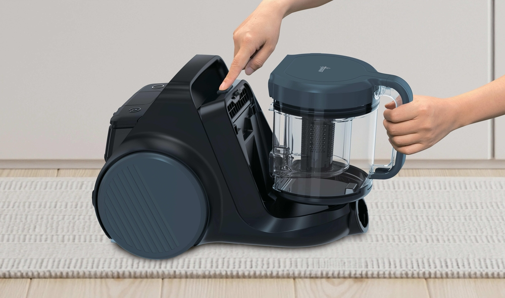 Eine Hand drückt einen Knopf an einem schwarzen Staubsauger, der auf einem Teppich steht.