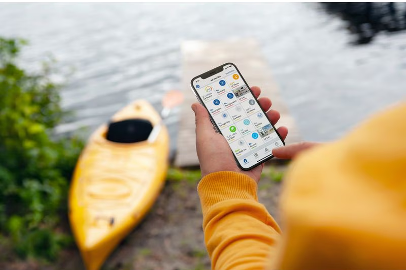 Person hält Smartphone mit App neben gelber Kajak am Wasser.